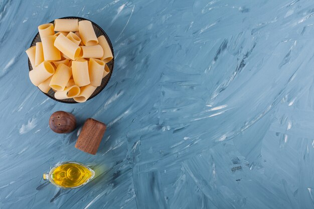 A bowl of dry raw tube pasta with oil and spices on a blue table .