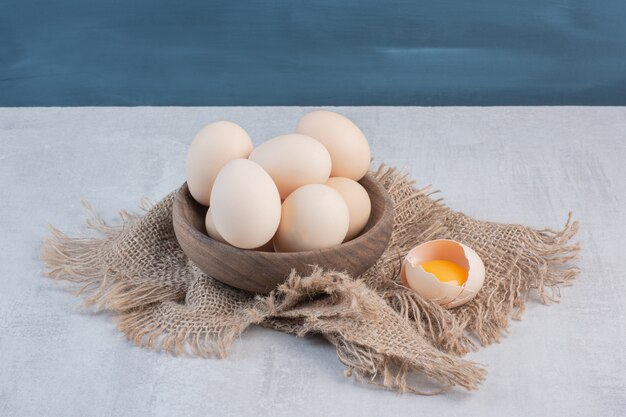 Bowl of eggs next to yolk in a shell on a piece of cloth on marble table.