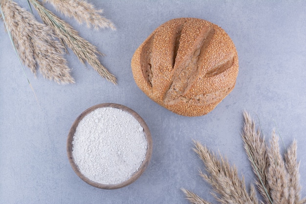 Bowl of flour loaf of bread and stalks of dried feather grass on marble surface