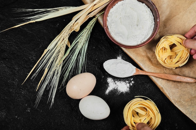 Bowl of flour, raw eggs, dry tagliatelle and wooden spoon on dark table.