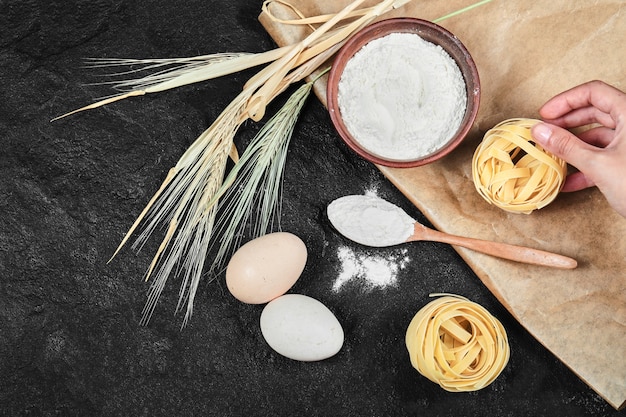 Bowl of flour, raw eggs, dry tagliatelle and wooden spoon on dark table.