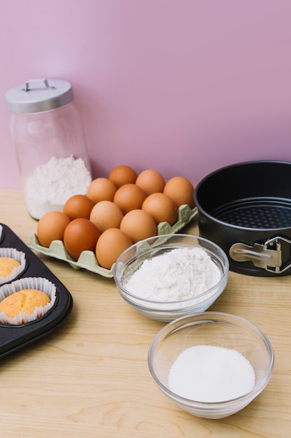 Free photo bowl of flour; sugar; eggs; cupcake and baking tin on wooden desk against pink background