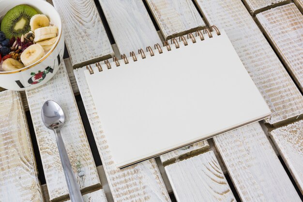 Bowl of fruit salad with spoon and blank spiral notebook on wooden textured background