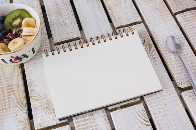 Bowl of fruits salad and blank spiral notepad on wooden pattern backdrop with spoon