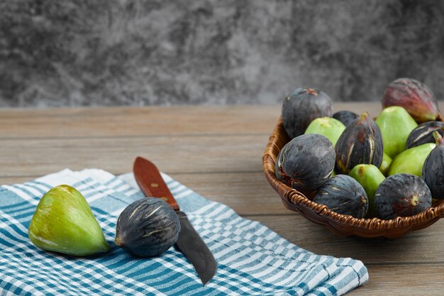 A bowl of green and black figs on wooden table with a knife and a tablecloth.