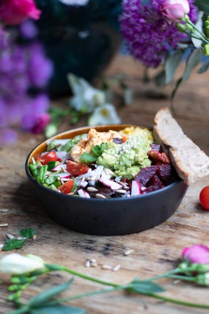 Bowl of healthy salad with avocado, vegetables, and seeds on wooden table among flowers