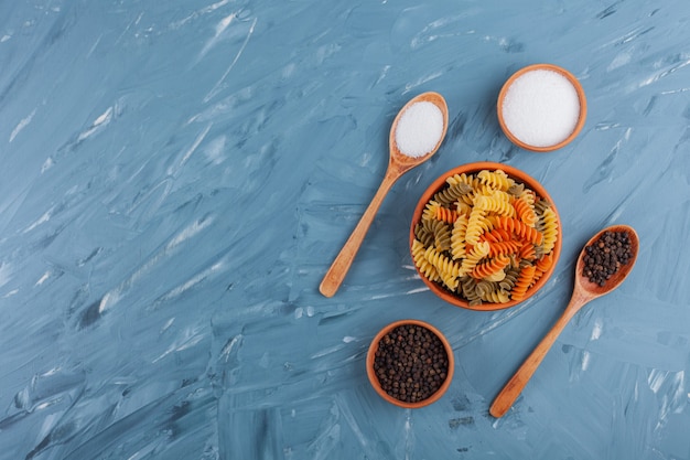 A bowl of multi colored raw spiral pasta with salt and pepper corns.