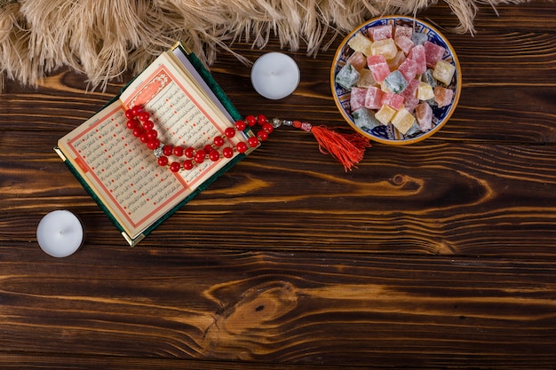 Bowl of multicolored lukum and red holy rosary beads and kuran with candles on wooden surface