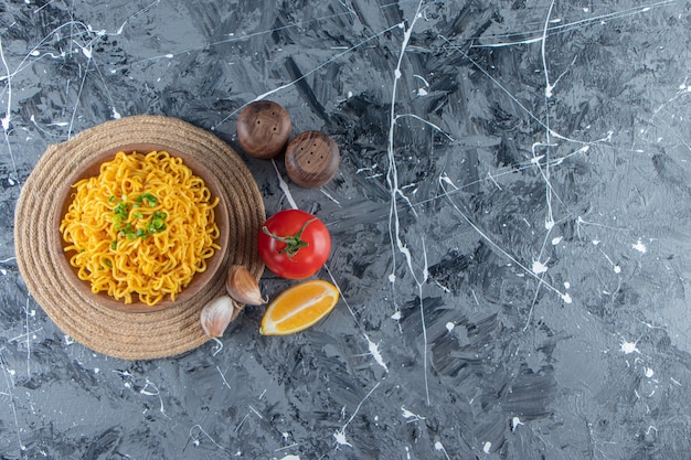 A bowl of noodle on a trivet next to tomatoes, lemon and garlic, on the marble background.