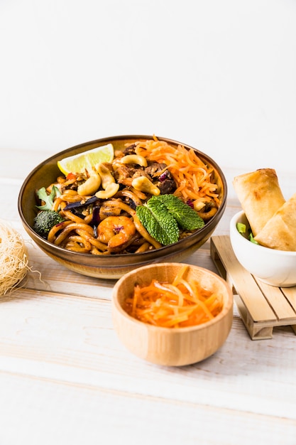 Bowl of noodles with spring roll and grated carrot on wooden table against white backdrop