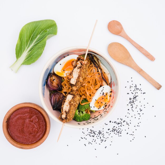 Bowl of noodles with vegetables and eggs with sauce; sesame seeds and wooden spoon on white backdrop