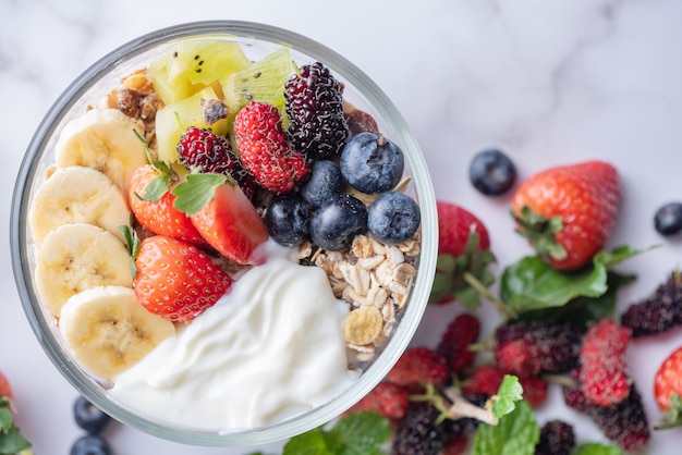 Bowl of oat granola with yogurt, fresh blueberries, mulberry, strawberries, kiwi, banana, mint and nuts board for healthy breakfast, top view, copy space, flat lay. vegetarian food concept.