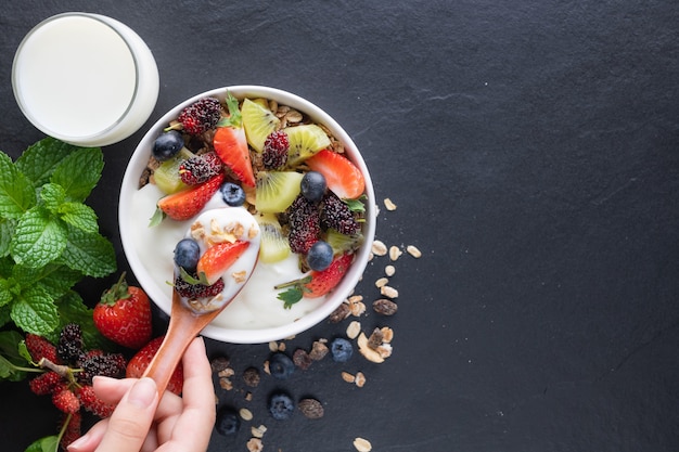 Bowl of oat granola with yogurt, fresh mulberry, strawberries, kiwi mint and nuts board for healthy breakfast, top view, copy space, flat lay. spoon in women's hands. vegetarian food concept.