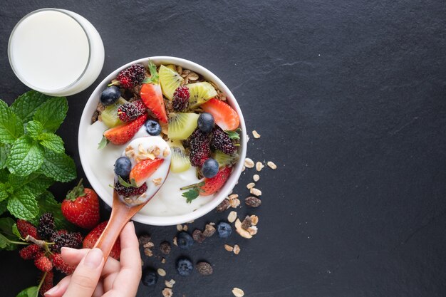 Bowl of oat granola with yogurt, fresh mulberry, strawberries, kiwi mint and nuts board for healthy breakfast, top view, copy space, flat lay. spoon in women's hands. vegetarian food concept.