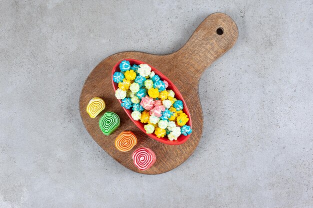 A bowl of popcorn candy next to marmelades on a wooden board on marble surface