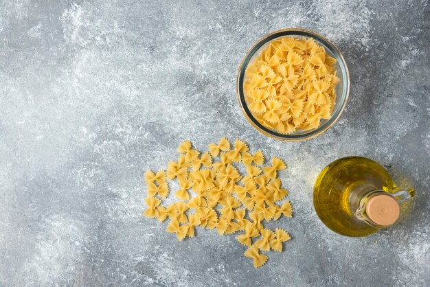 Bowl of raw farfalle pasta with bottle of olive oil on marble background. 