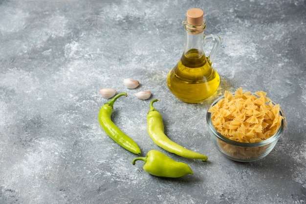 大理石の背景にオリーブオイルと野菜のボトルと生のファルファッレパスタのボウル。