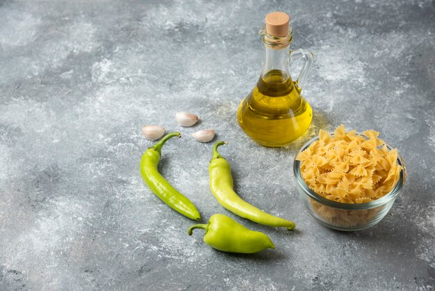 Bowl of raw farfalle pasta with bottle of olive oil and vegetables on marble background. 