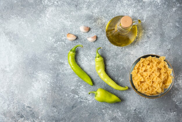 Bowl of raw farfalle pasta with bottle of olive oil and vegetables on marble background. 