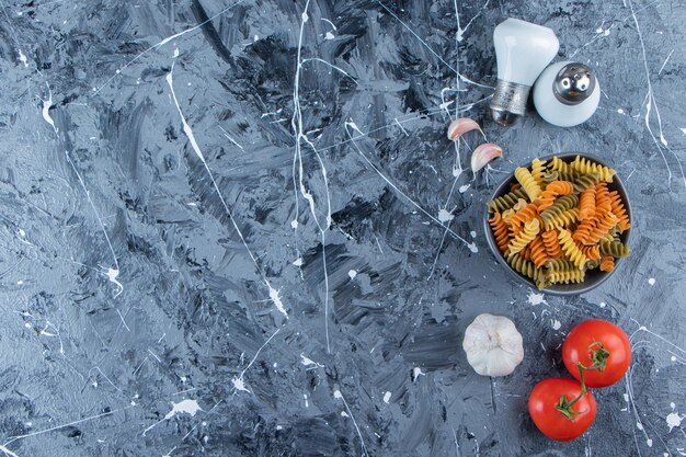 A bowl of raw pasta with fresh vegetables and spices on a marble background.