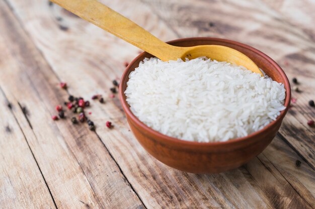 Bowl of raw white rice in wooden bowl near black pepper over wooden plank
