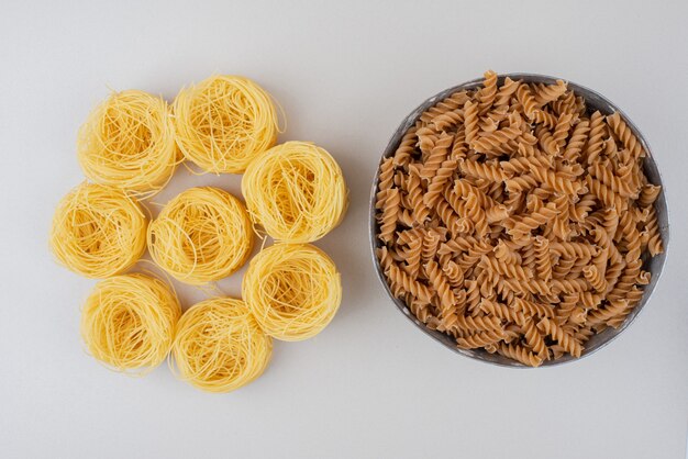 Bowl of spiral pasta and spaghetti nests on white surface.