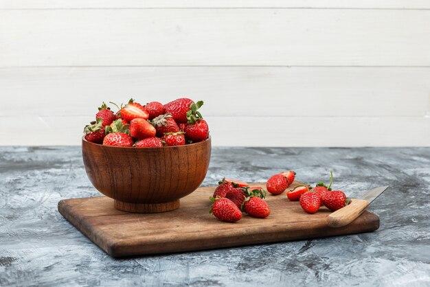 A bowl of strawberries and a knife on a wooden cutting board on a dark blue marble and white wooden board background. close-up. free space for your text