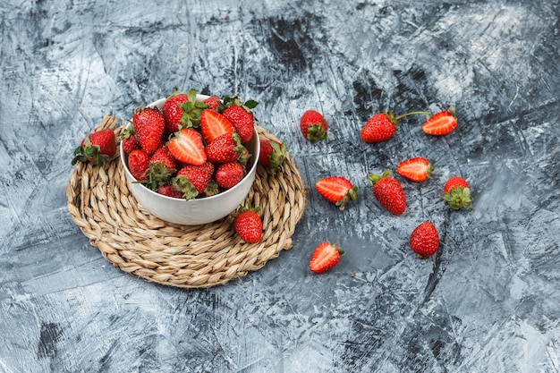 A bowl of strawberries on a round wicker placemat on a dark blue marble background. .