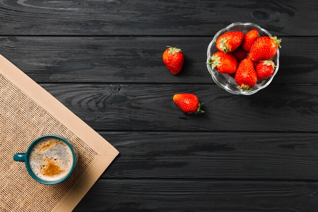 Bowl of strawberry and coffee cup on jute placemat over black textured surface