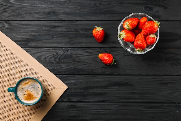 Bowl of strawberry and coffee cup on jute placemat over black textured surface