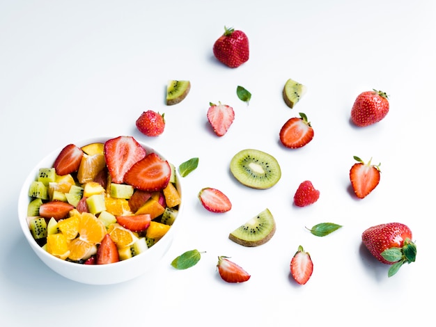 Чаша вкусного фруктового салата возле кусочков клубники и киви на белом фоне