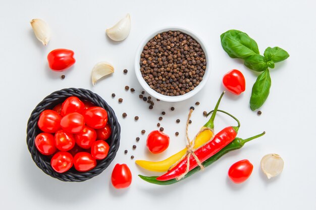 A bowl of tomatoes with a bowl of black pepper, garlic, leaves, chili pepper top view on a white surface