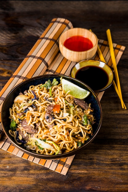 Bowl of udon noodles with soya and red chili sauces on placemat against wooden desk