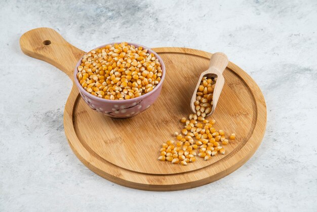 Bowl of uncooked corn grains with spoon on wooden board.