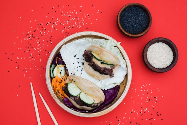 Bowl of white and black sesame seeds with gua bao in steamer on red backdrop