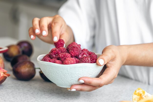 A bowl with raspberries in female hands on a white kitchen table