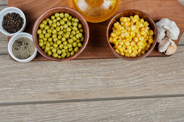 Bowls of boiled sweet corn and green peas, spices, oil, and vegetables on a wooden board.