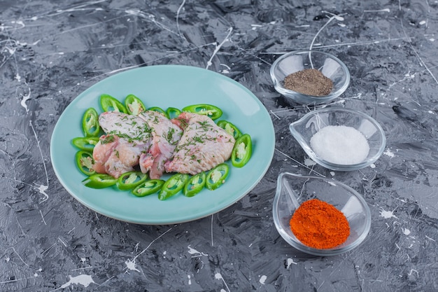 Bowls full of different spices next to plate with wings and peppers on the blue surface