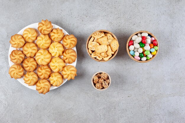 Bowls of glazed peanuts, candies and biscuit chips next to a plate of cookies on marble surface.