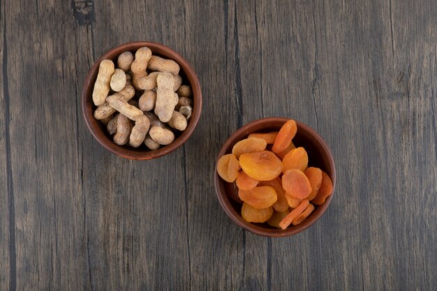 Bowls of healthy dried apricot fruits and peanuts in shell on a wooden table .