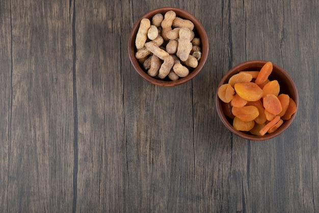 Bowls of healthy dried apricot fruits and peanuts in shell on a wooden table .