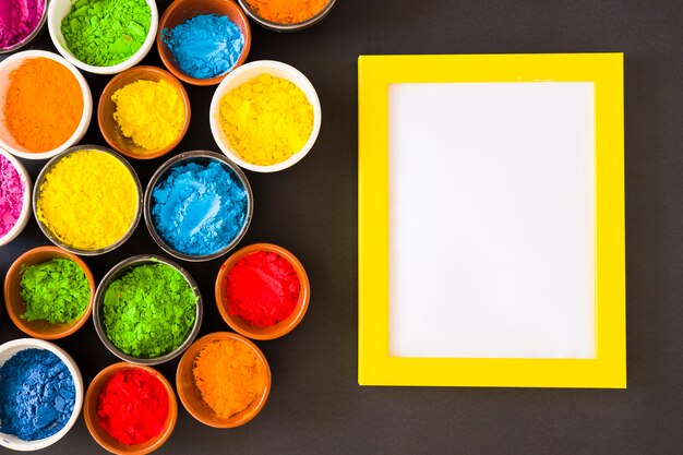 Bowls of holi color powder near the white frame with yellow border on black background