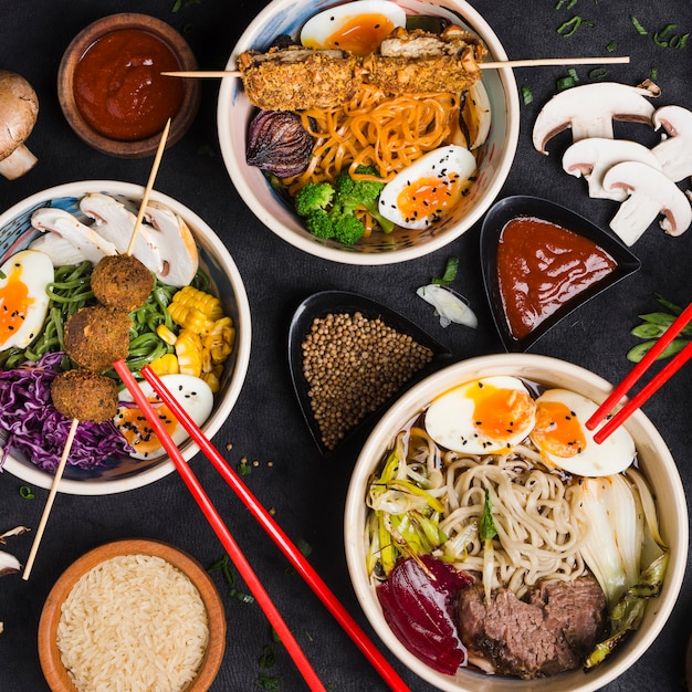 Bowls of noodles with eggs; vegetables and sauces on black background