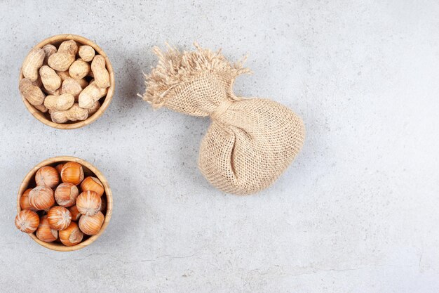 Bowls of peanuts and hazelnuts next to a sack on marble background. High quality photo