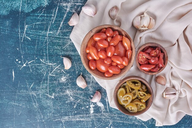 Bowls of pickled tomatoes and jalapenos on blue with garlic and tablecloth. Top view. 