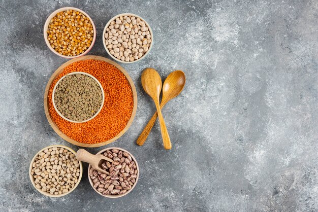 Bowls of various uncooked beans, lentils and corns on marble table surface.