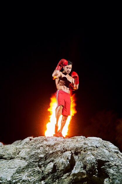 Foto gratuita campione di pugilato che posa sopra una roccia con fuoco che brucia sul