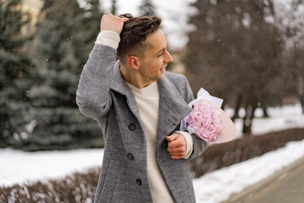 Boy friend with a bouquet of pink flowers hydrangea waiting for his girl friend outdoors while snow is falling. Valetnine`s day concept, wedding proposal. 
man goes on a date.