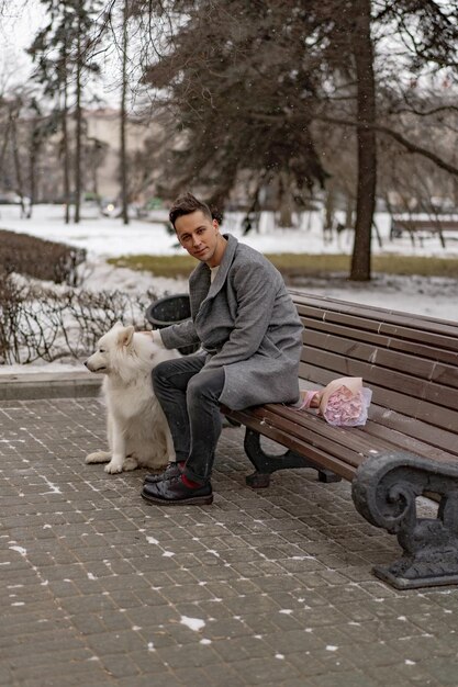 ピンクの花の花束を持ったボーイフレンドは、彼のガールフレンドを待っていて、犬と一緒に歩いたり遊んだりしています。雪が降っている間、屋外で。 Valetnineの日のコンセプト、プロポーズ。マンゴー