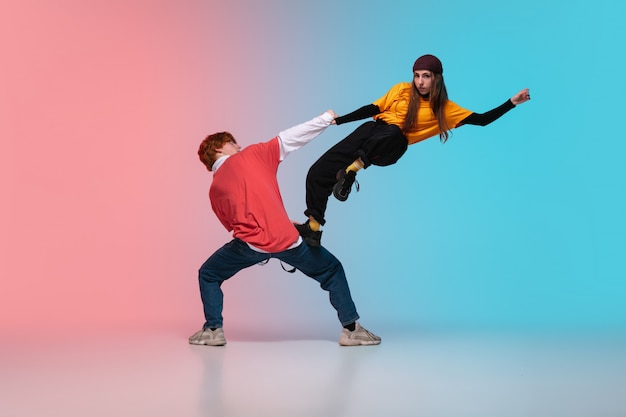 Free photo boy and girl dancing hip-hop in stylish clothes on gradient background at dance hall in neon light.